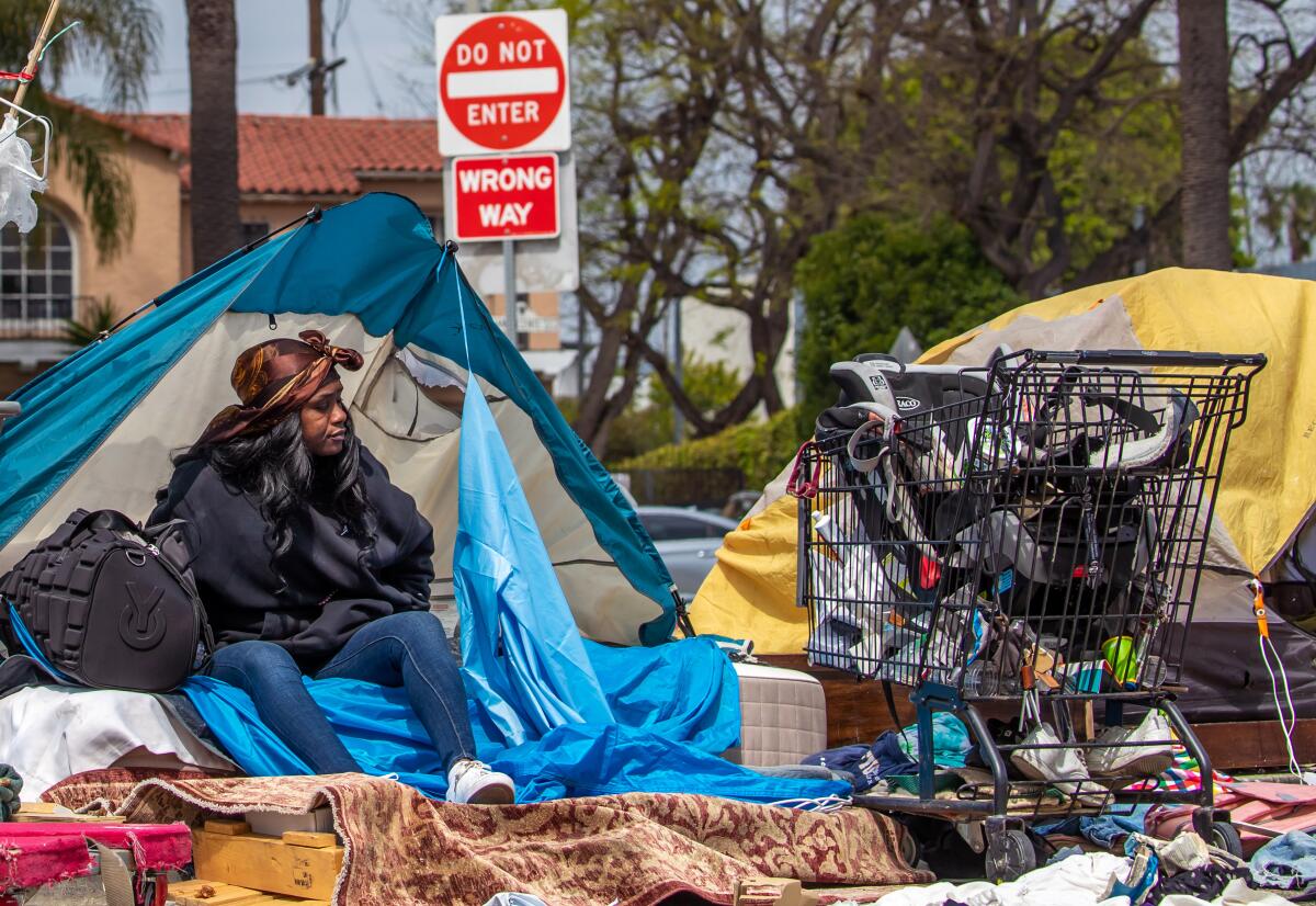 A woman sits in the opening of a tent pitched on a sidewalk next to a shopping cart filled with her possessions.