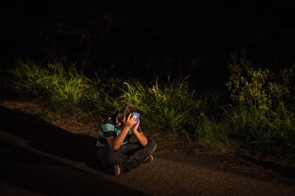 A boy sits cross-legged on the ground at night while shielding his face from a light.