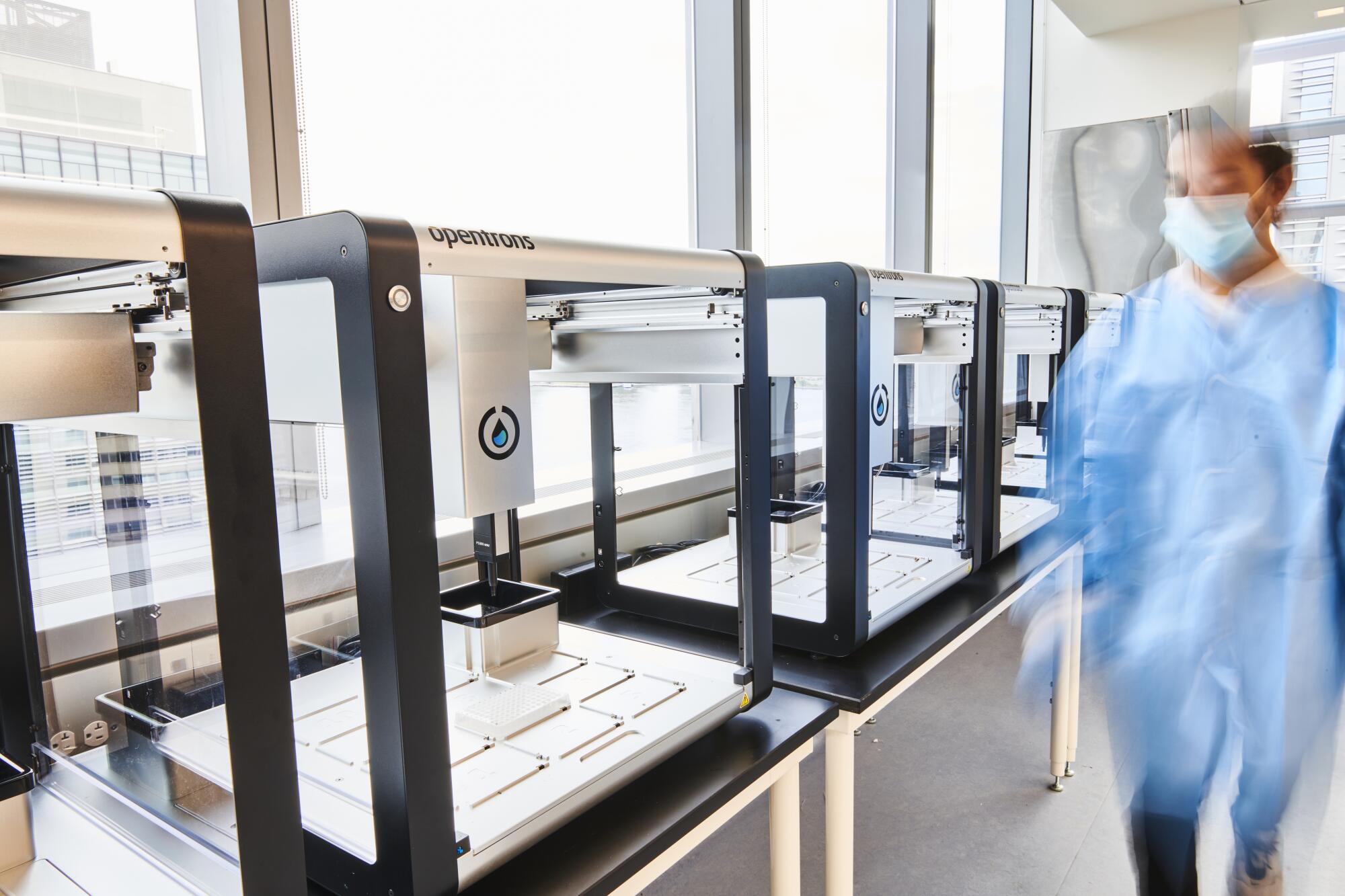 A masked person in a blue lab coat stands near aquarium-like machines on a table