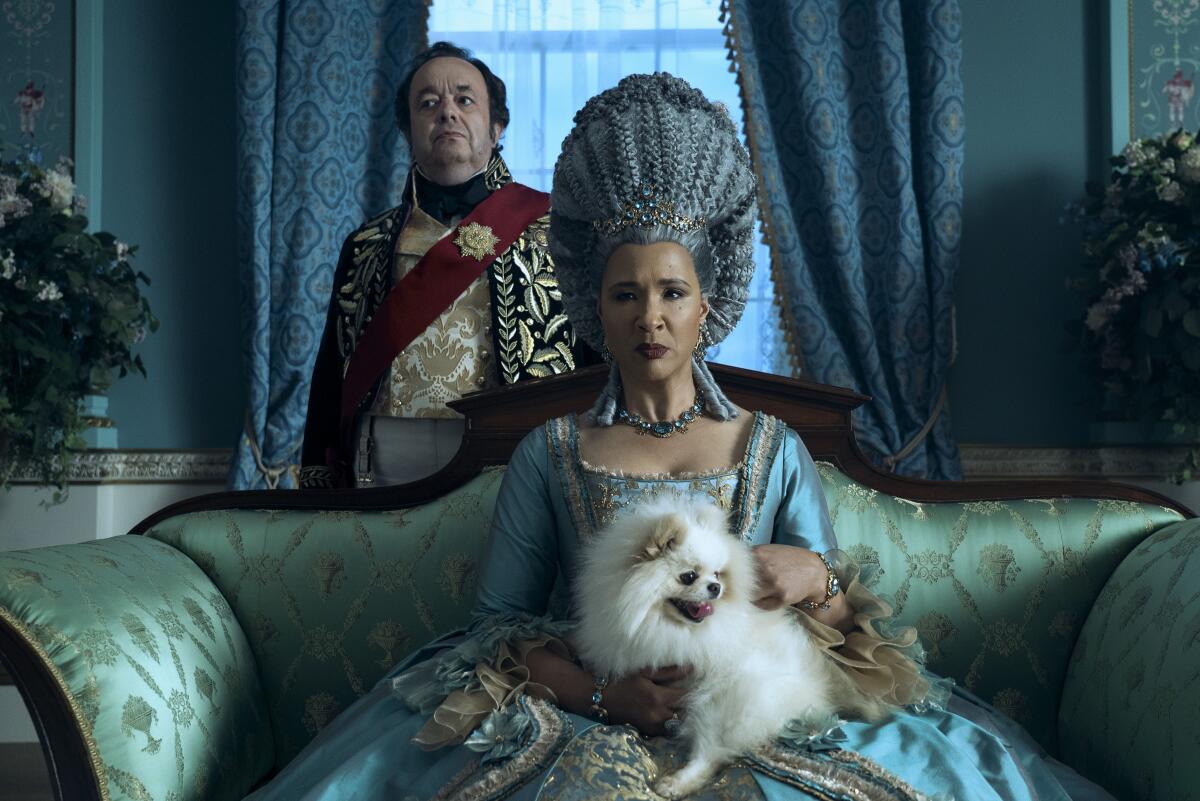 Brimsley stands behind Queen Charlotte, who is sitting on a couch with a dog on her lap.