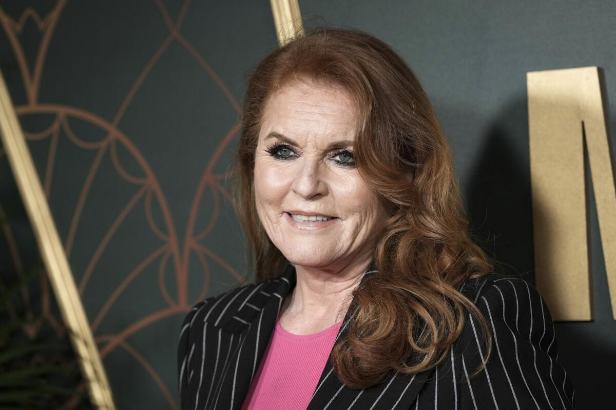 Sarah Ferguson, the Duchess of York, smiles in a pinstripe blazer and pink shirt against a black-and-gold background.
