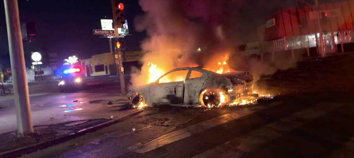 Flames engulf a car on a city street after dark.