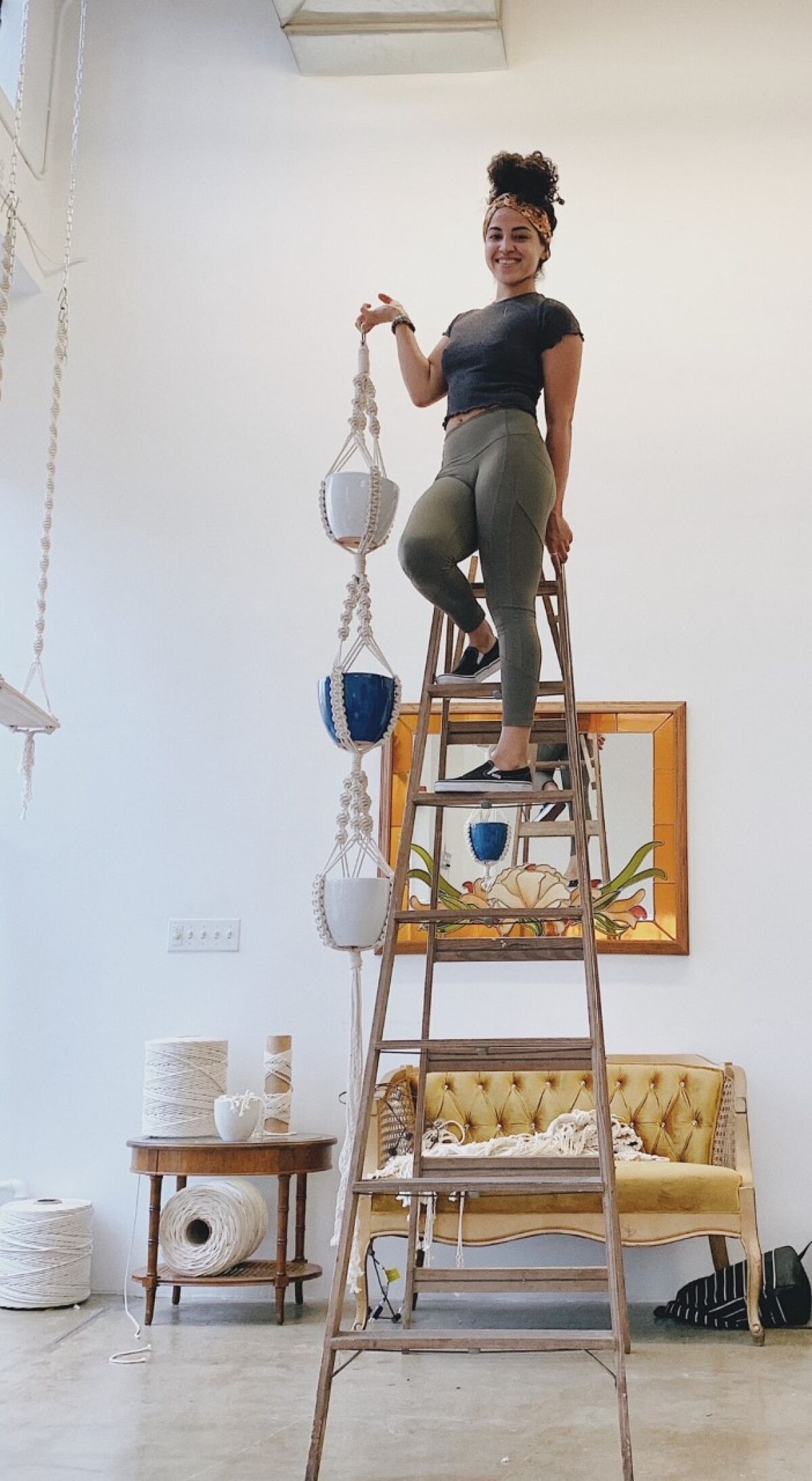Lola Morales stands on a ladder holding a macramé plant hanger that holds three potted plants.