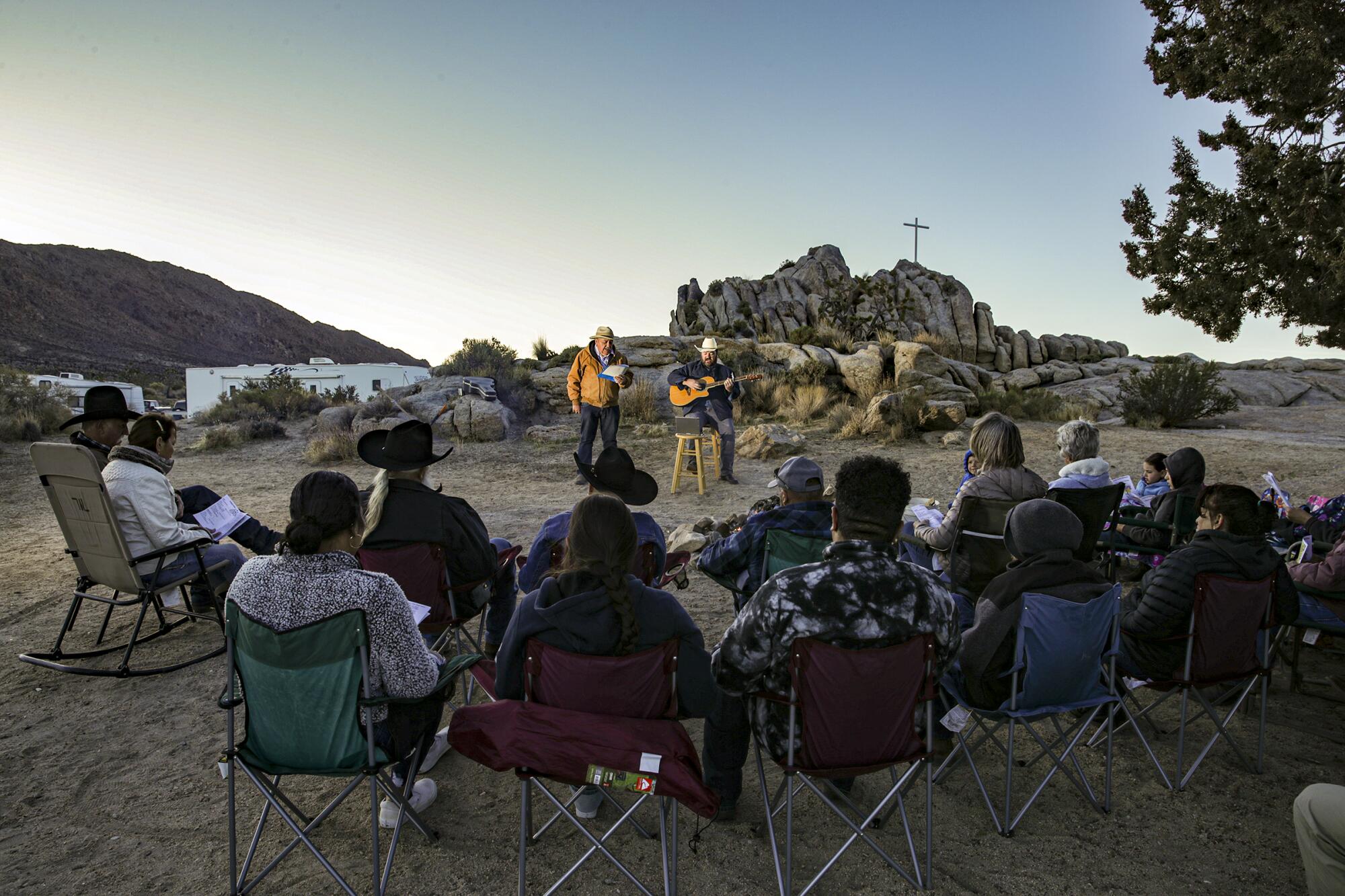 People sit in folding chairs, watching a preacher and a man with a guitar in front of a rock formation topped with a cross