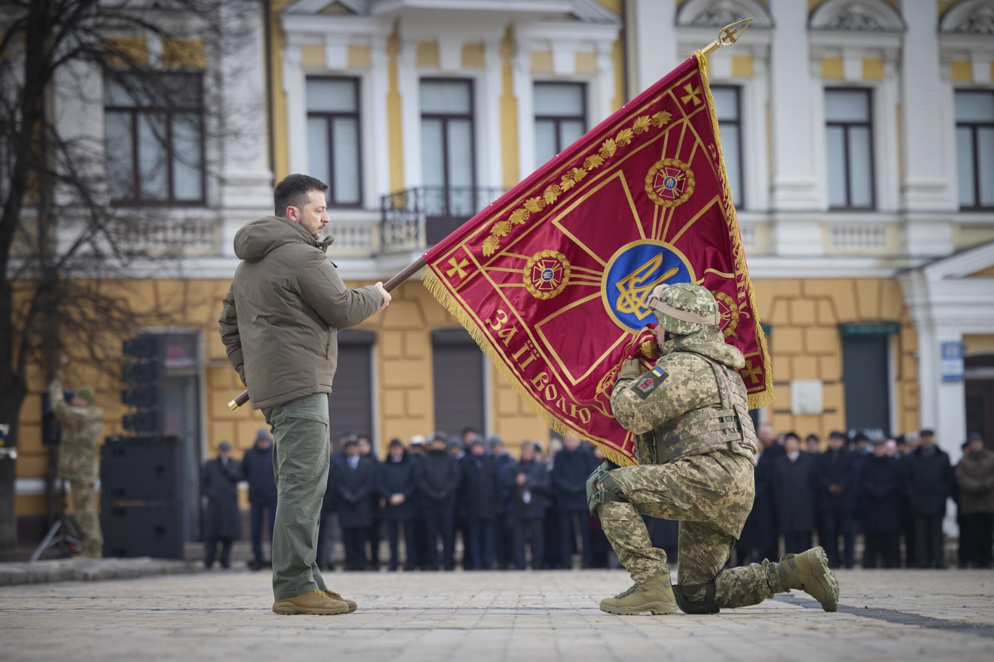 In a public square, Ukrainian President Volodymyr Zelensky holds the flag of a military unit as a kneeling officer kisses it.
