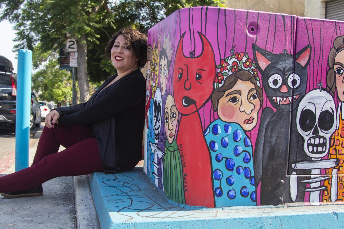 Artist Yvette Roman sits next to her art, a painting of various puppet faces on a utility box in Sherman Heights