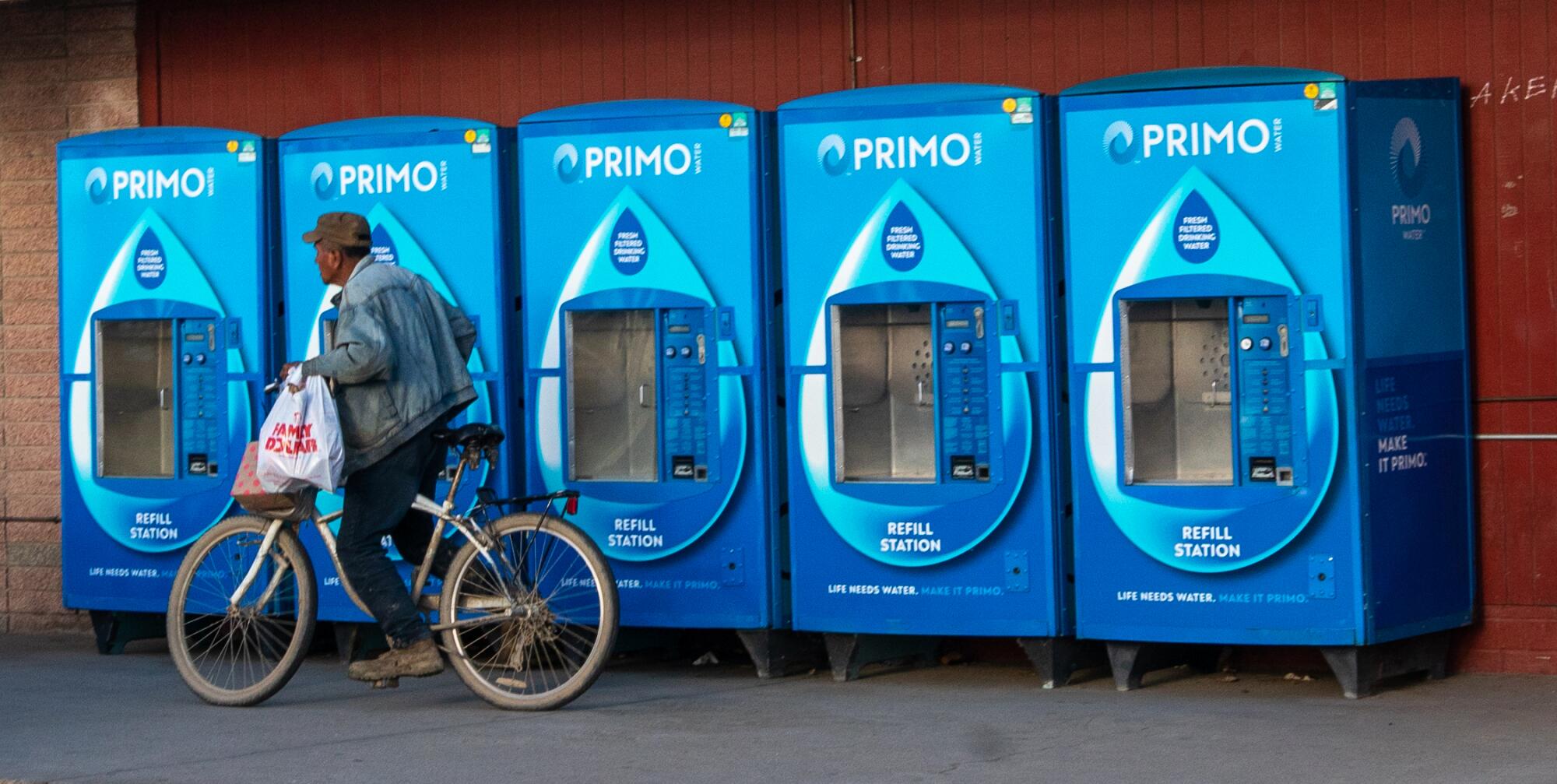 A man pedals a bicycle past a row of water-dispensing machines.  
