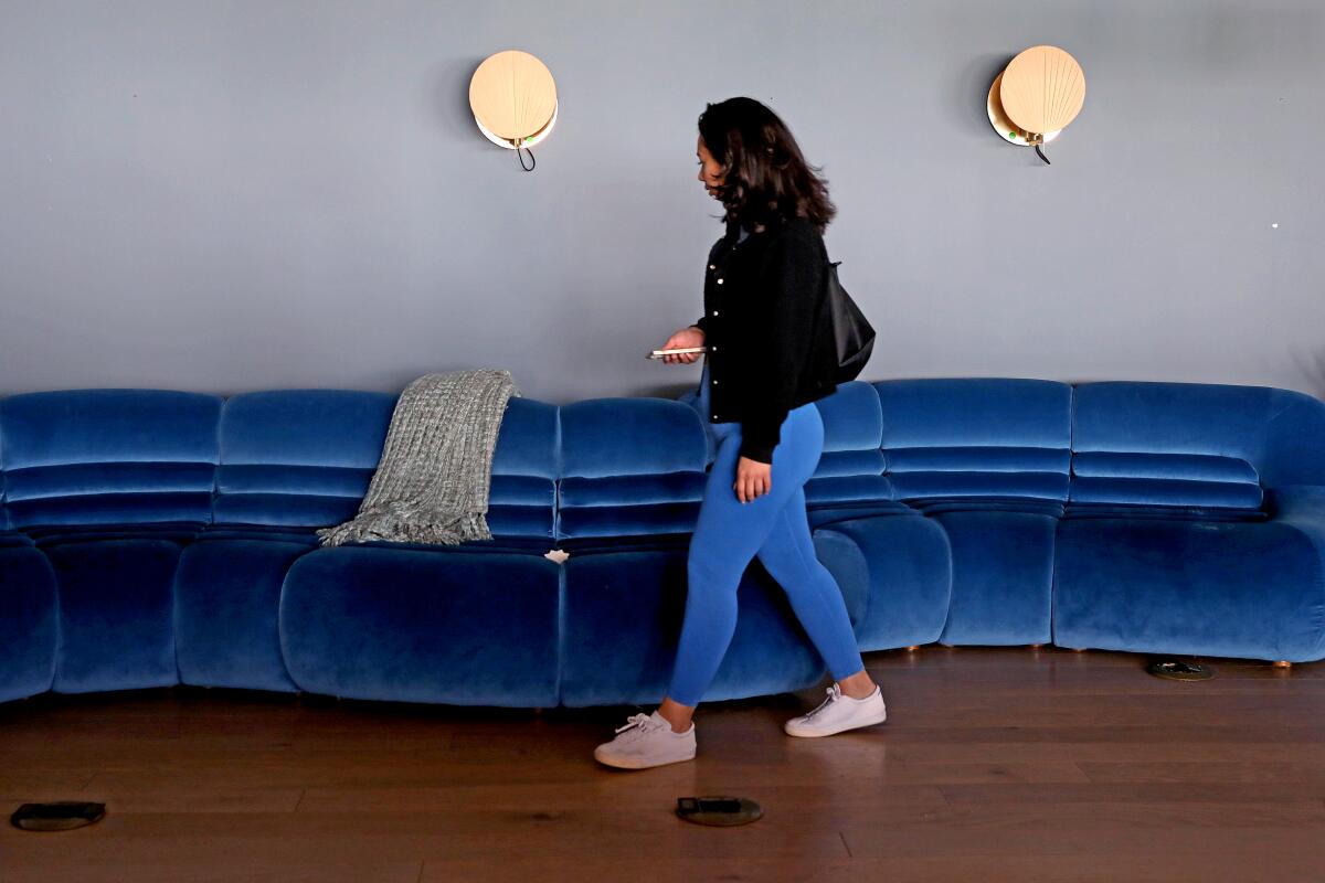 A dark-haired woman in a black jacket and blue leggings walks past a blue couch at the Wing.