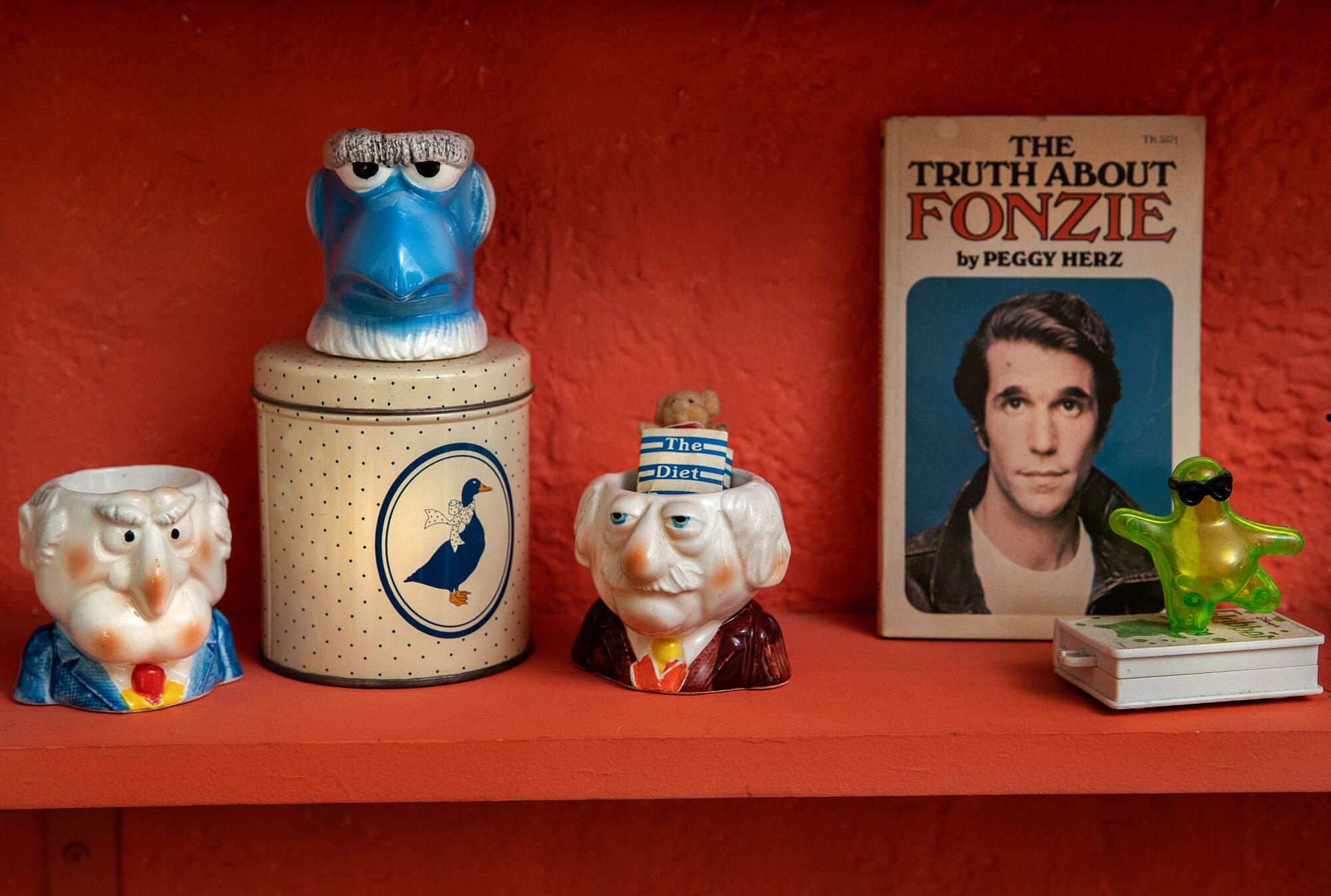 Items on an orange shelf, including a book on Fonzie, a plastic flubber toy and muppet-themed mini vases.
