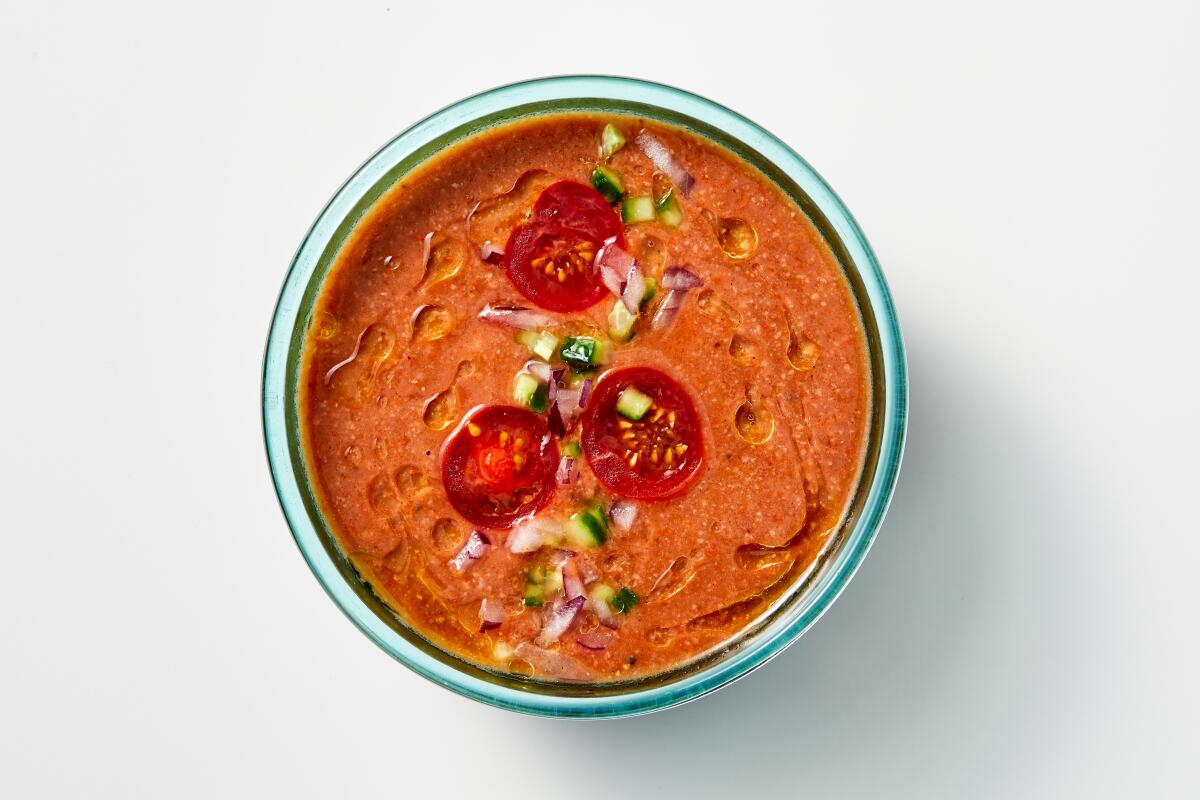 Overhead view of a bowl with reddish soup. On top are drips of olive oil, sliced tomatoes and chopped cucumber.