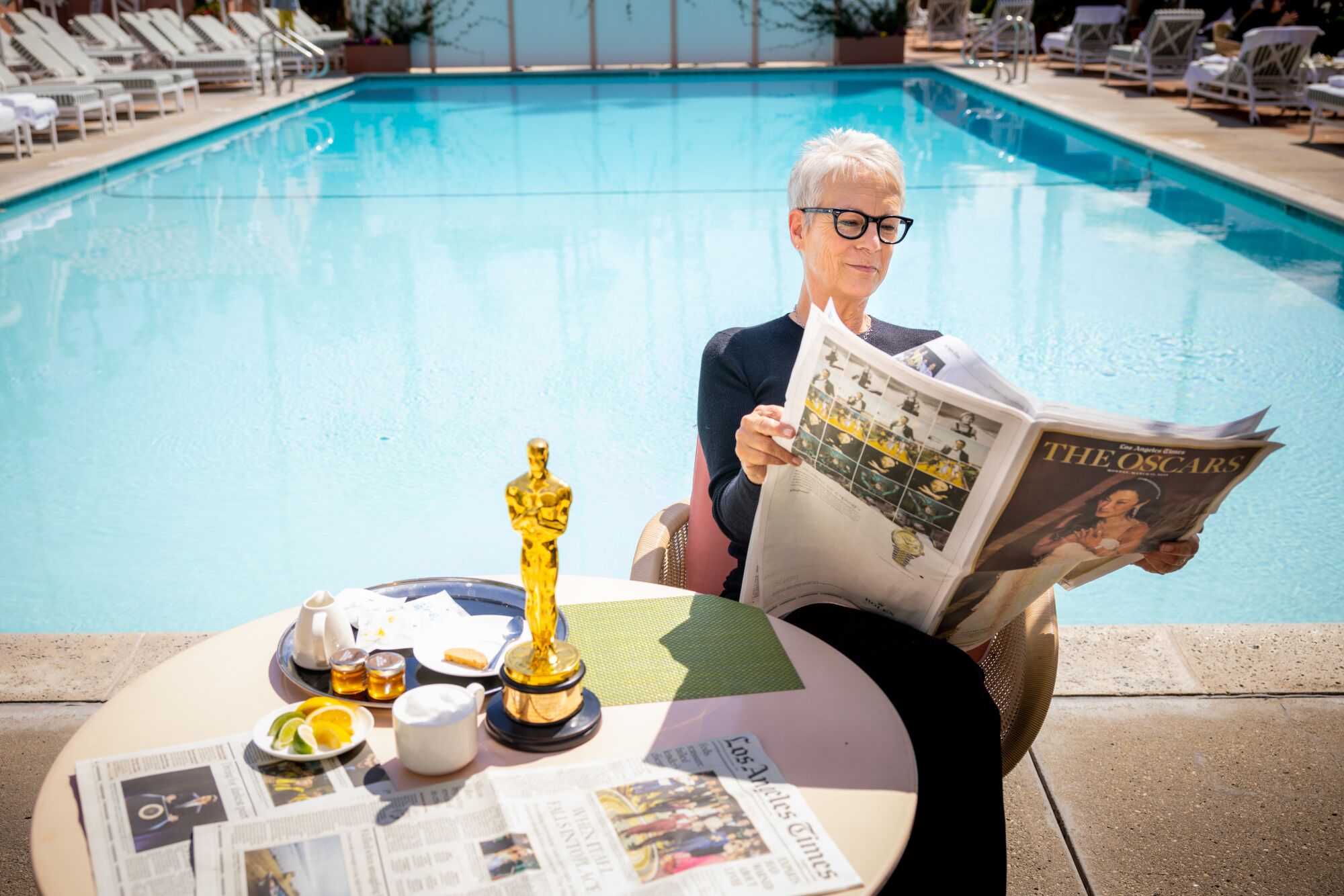A woman with glasses looks at a newspaper section.  Her Oscar is on the table.
