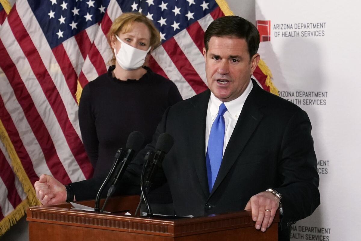 Arizona Gov. Doug Ducey speaks emphatically at a lectern as a masked person stands behind him.
