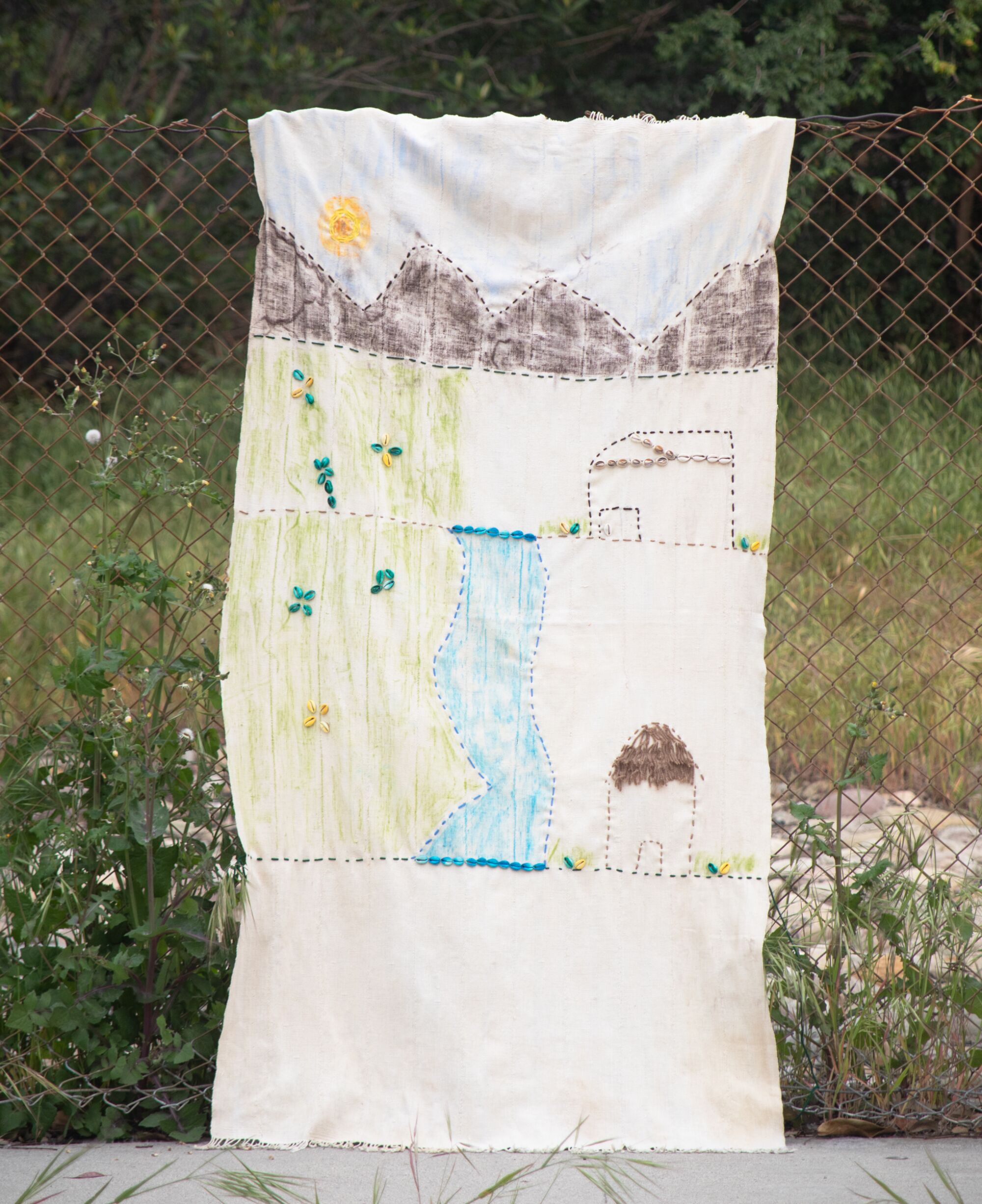 A textile depicting a village in the mountains hangs on a fence by the 118 freeeway