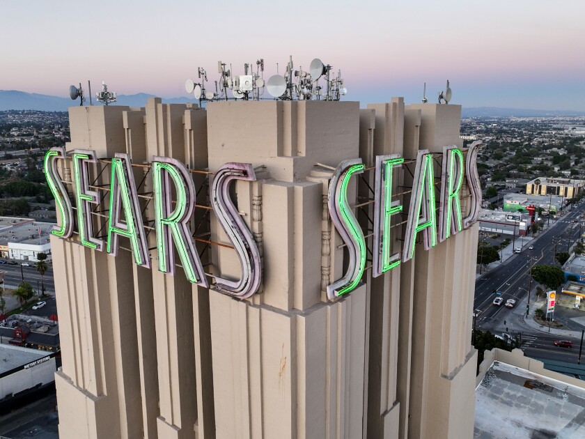 Closeup of the top of a multistory retail building with "Sears" in neon green on either side that overlooks a city.