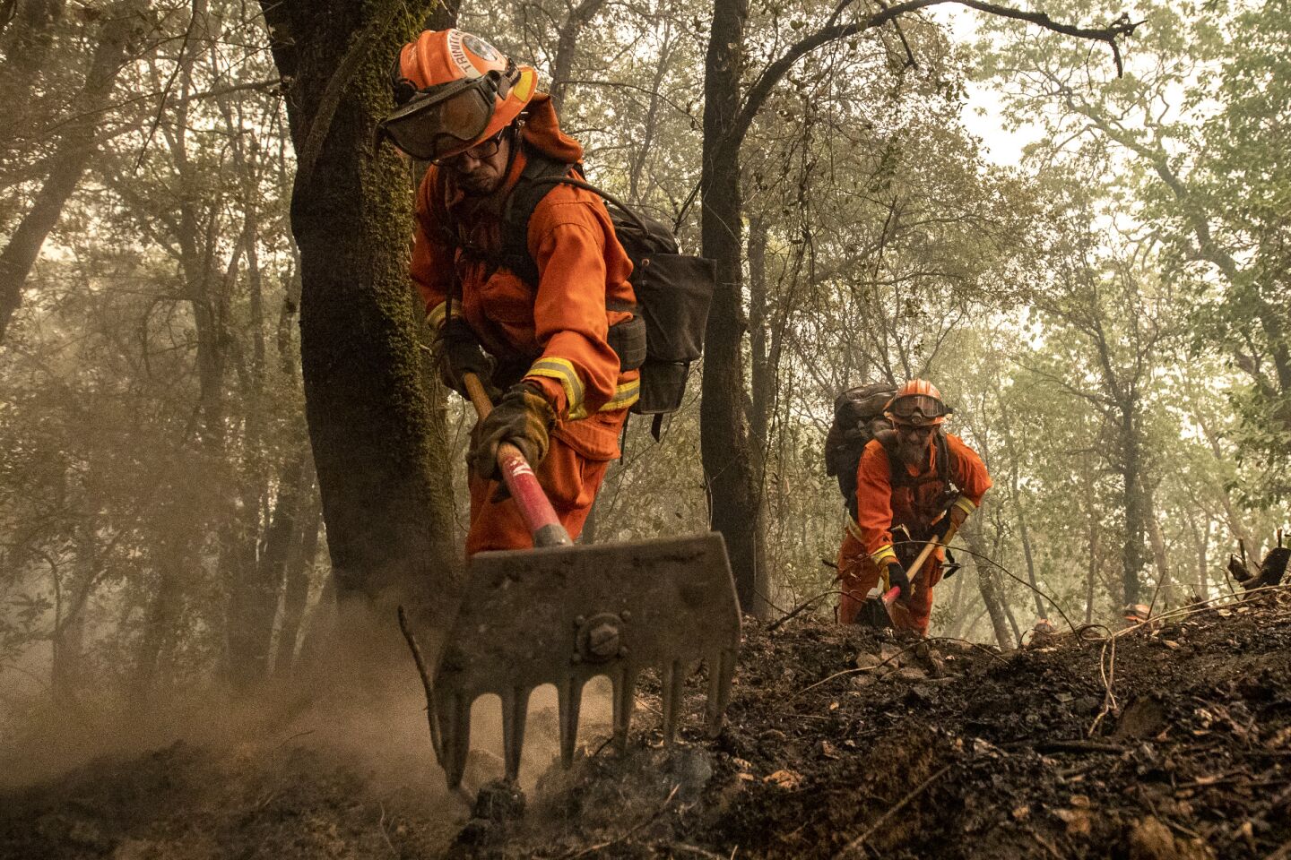 Two men wearing orange suits use hand tools to dig into the earth after a fire burned through.