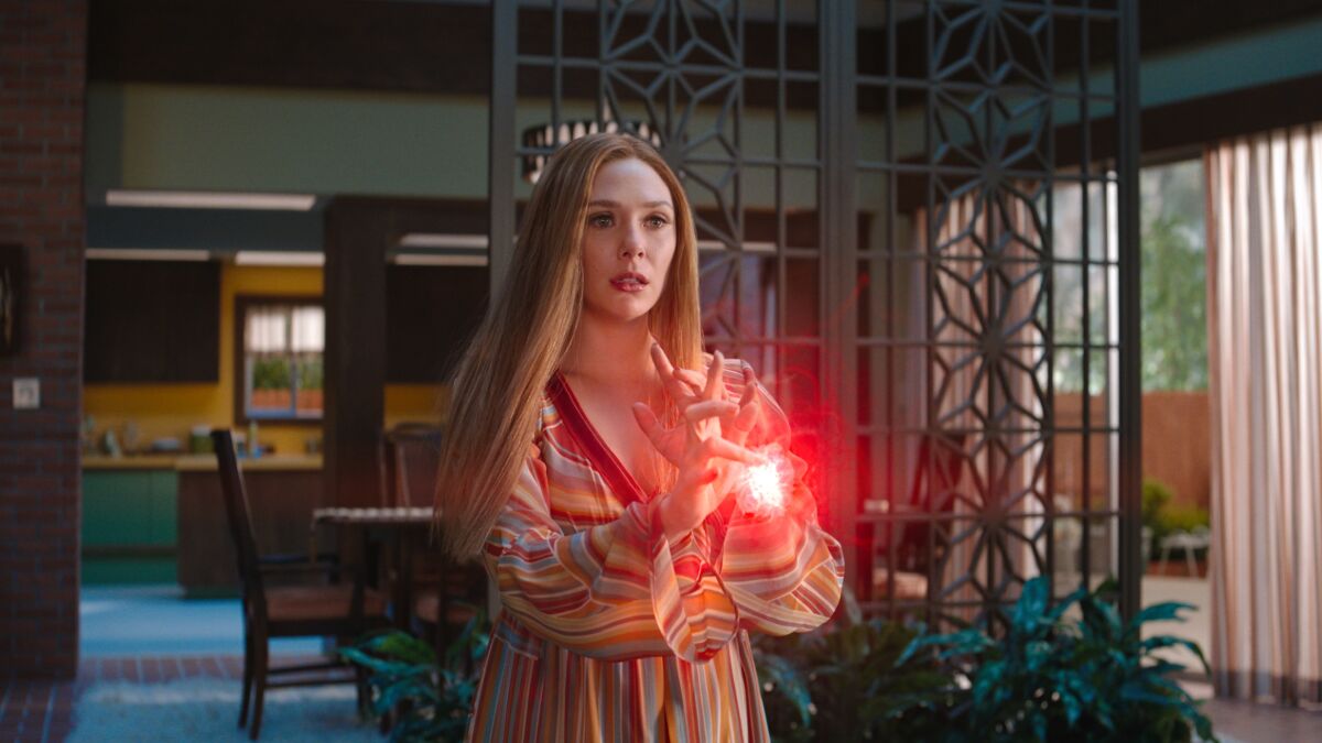 Elizabeth Olsen, in a striped ’70s-era dress on a period set, holds her hands in front of her as fire forms between them.