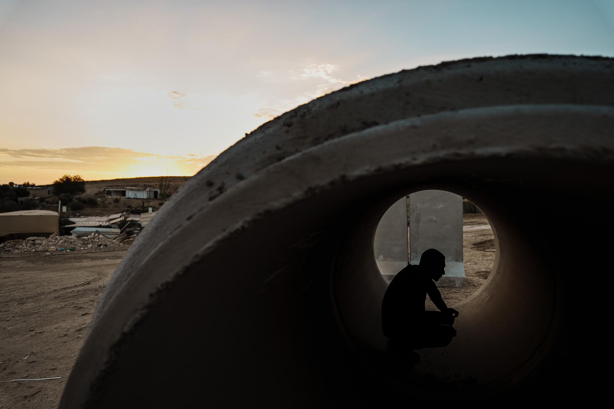 A person, seen in silhouette, sits inside a huge pipe in a desert setting 