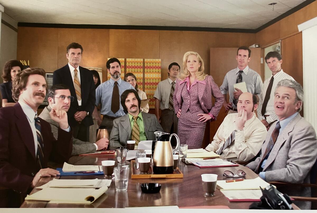 A scene from the 2004 comedy "Anchorman" with many people seated or standing around a table.