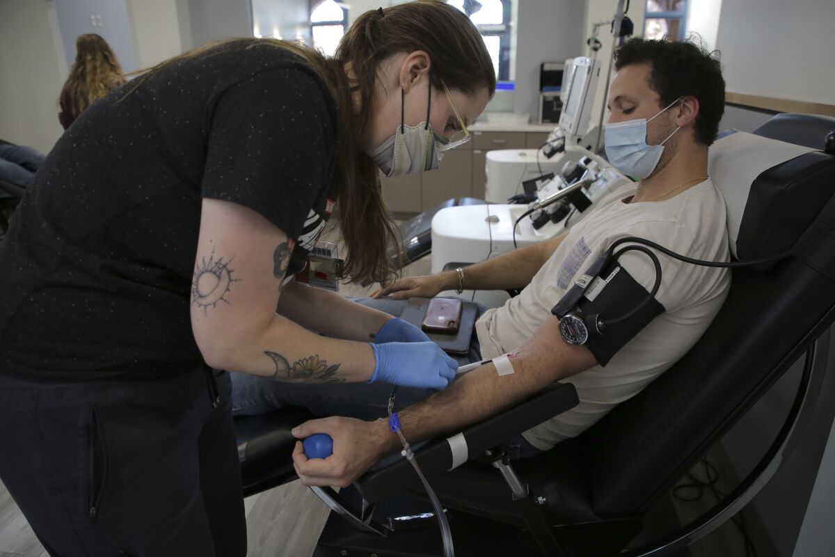 A man sitting in an easy chair has a needle inserted in his arm by a technician at a blood donation center