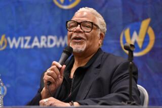 Ken Page sits at a table and wears thick-framed eyeglasses and a black suit while speaking into a microphone