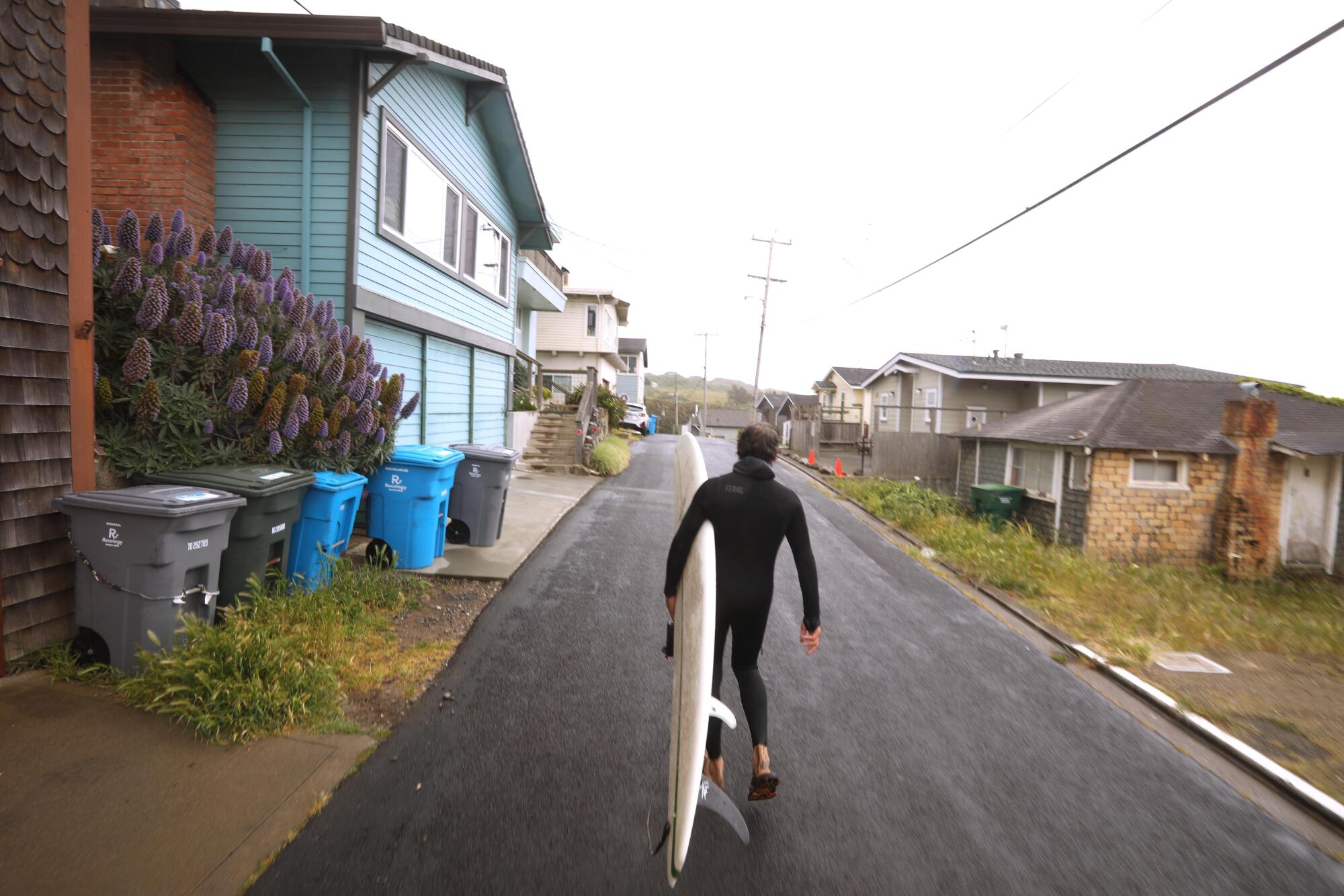 A man in a wetsuit carries a surfboard down a narrow street.