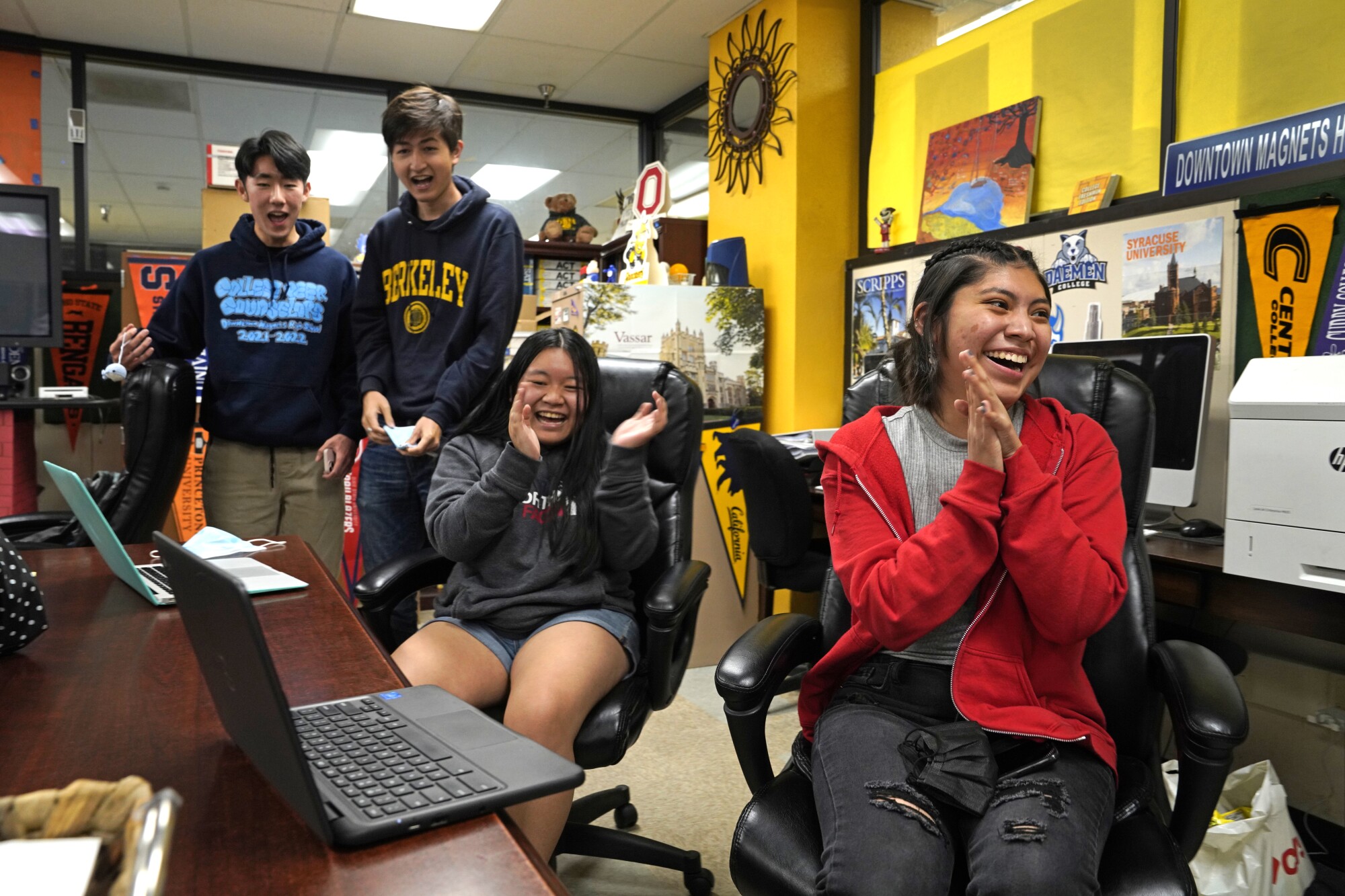 Two female students smile and clap while two male students standing nearby smile.