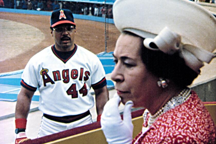 A baseball player in an Angels uniform staring at a woman in a large hat, white gloves and a red dress