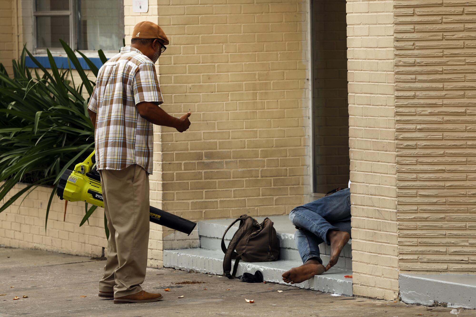 A man in a plaid shirt and khaki pants holding a leaf blower speaks to a person lying on steps near a backpack 