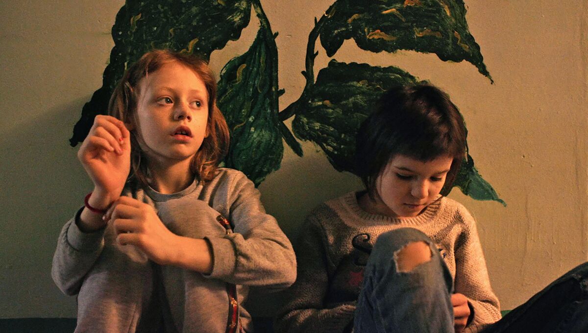 Two girls lean against a wall painted with images of plants in a scene from "A House Made of Splinters." 
