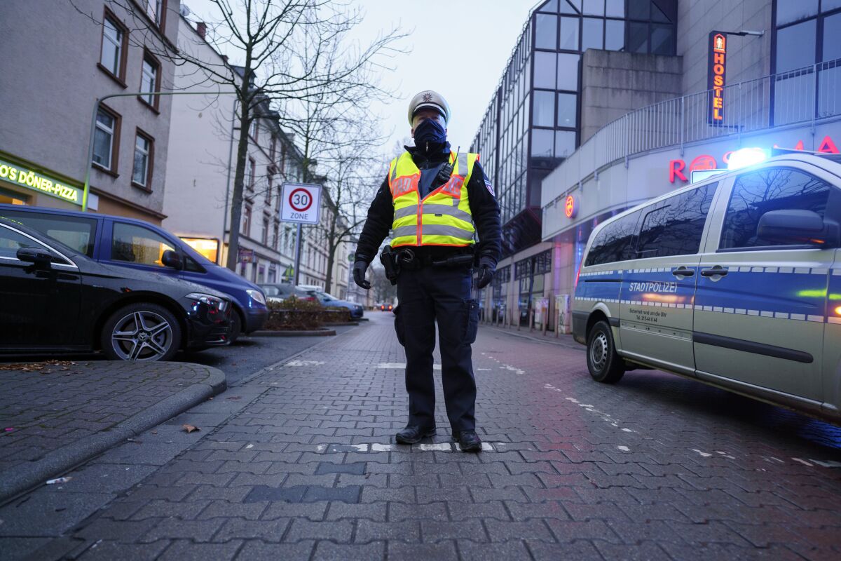 A masked policeman stands in the middle of a street flanked by parked cars and buildings.