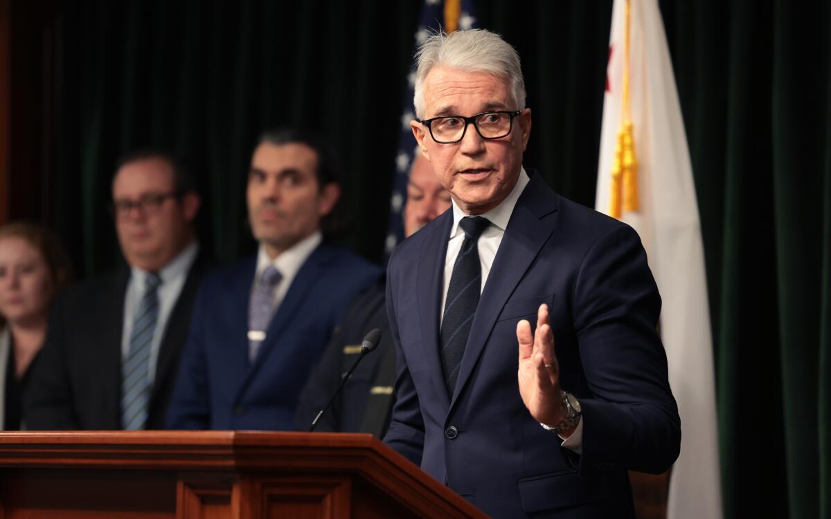 A man with gray hair, wearing glasses and dark suit and tie, gestures while speaking at a lectern near other people 
