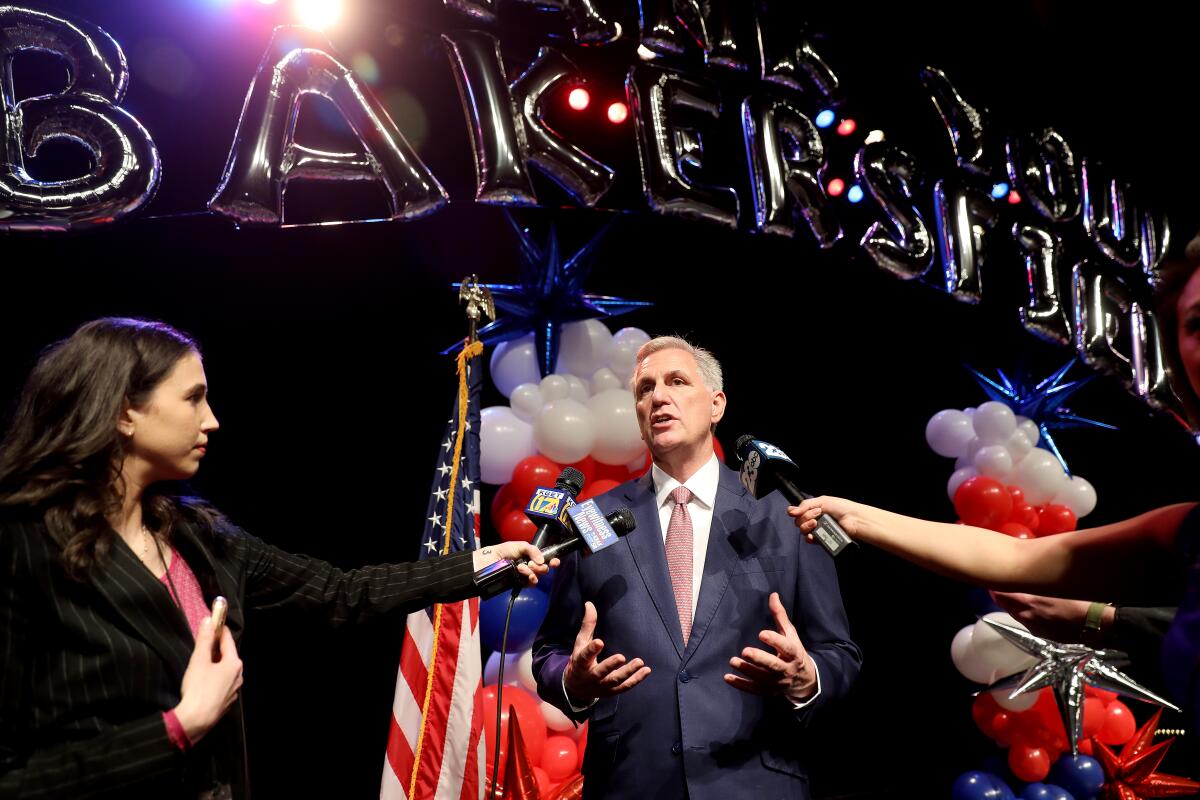 Large balloons spell "Bakersfield" above Kevin McCarthy as he speaks into mikes by red, white and blue decor and a U.S. flag