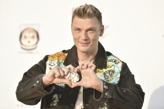 A man with short blond hair wearing a jean jacket and forming a heart shape with his hands