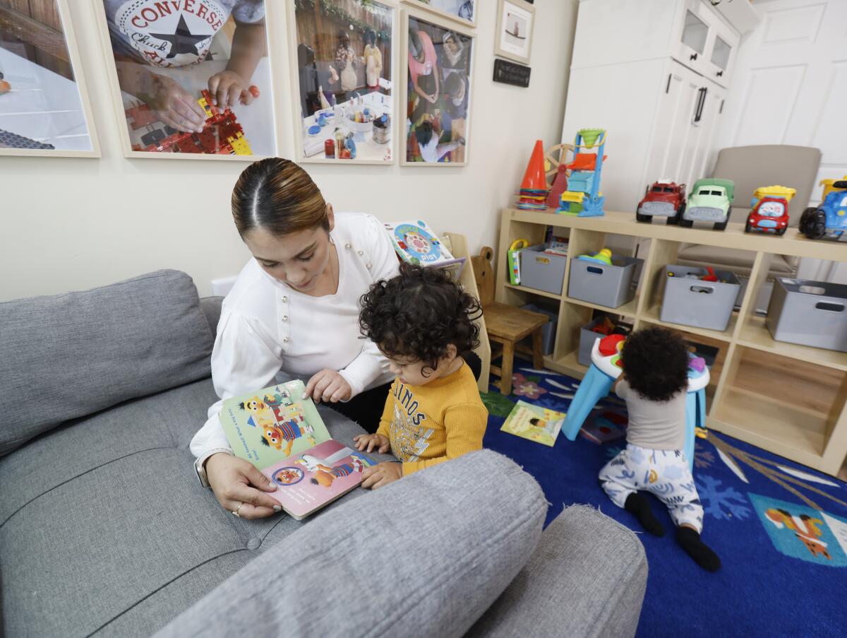A childcare assistant, turns the pages as she reads to 20-month-old boy, while an 8-month-old plays behind them.