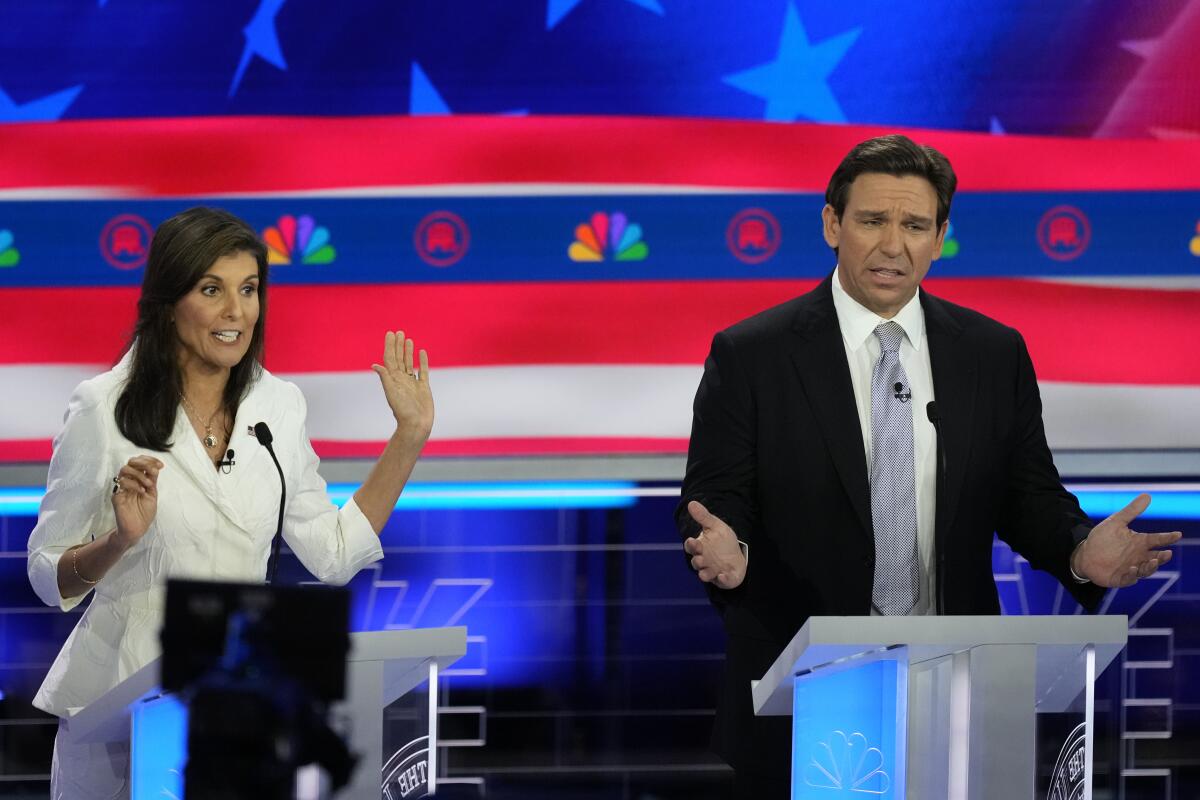 A woman in a white suit and a man in a black suit and gray tie gesture as they speak on stage.