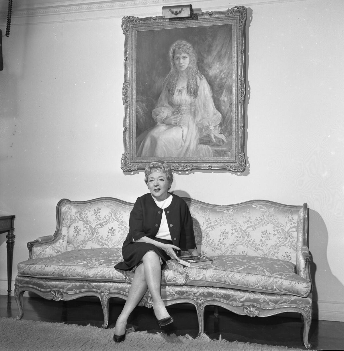 A woman sits on a couch beneath a large framed portrait of a young woman with long curls.