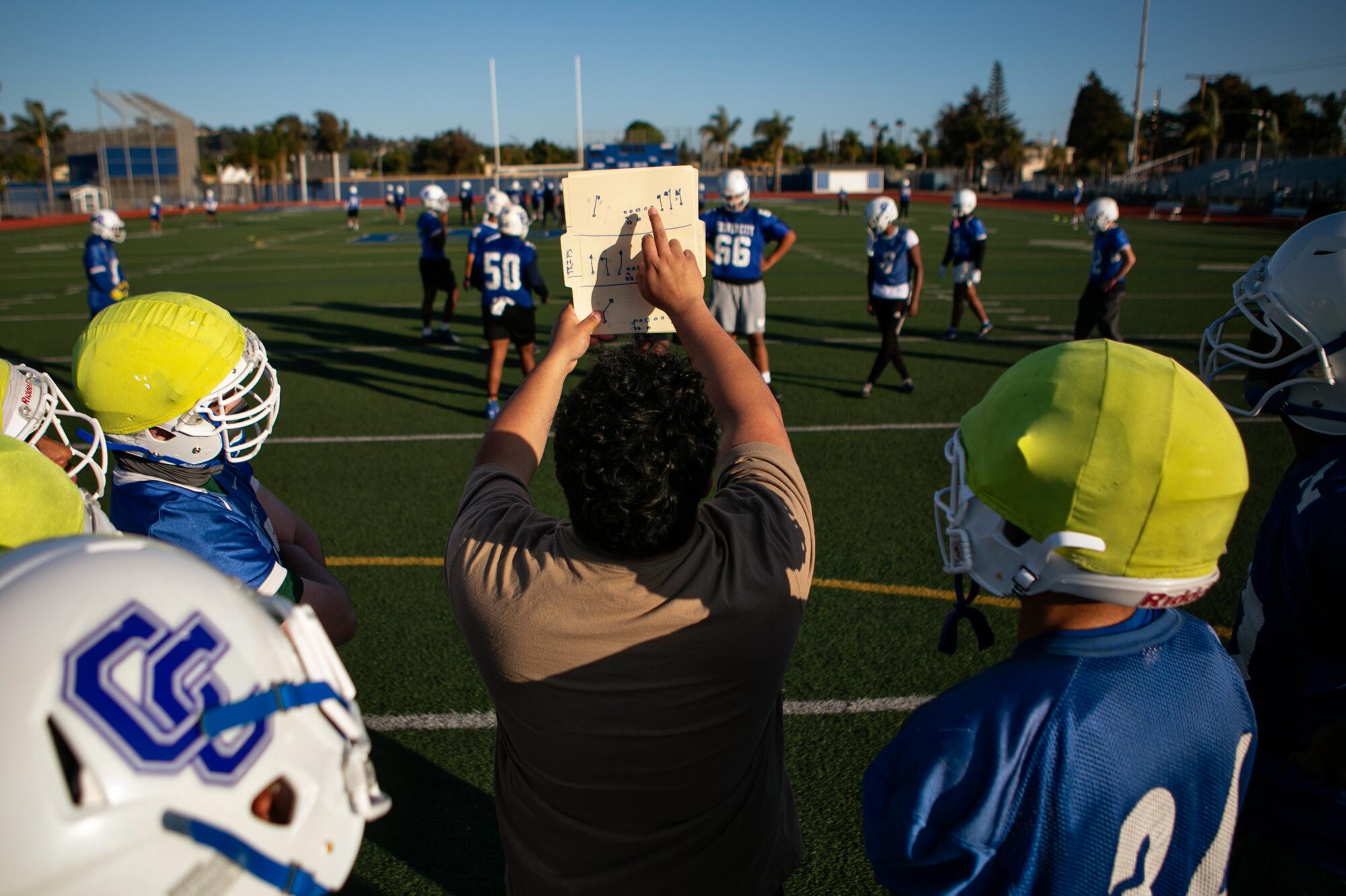 A coach, alongside players on the sidelines, holds up and points to a playbook.