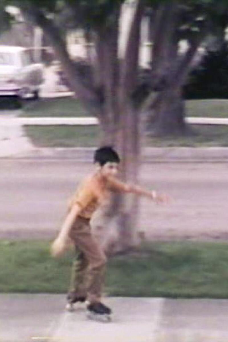 An old photo of a boy roller-skating past a young tree in a neighborhood.