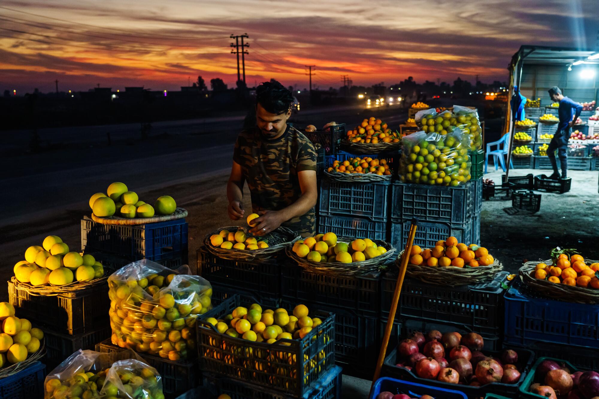 A man hold a piece of fruit as he stands amid produce piled in crates, baskets and plastic bags along the side of a highway
