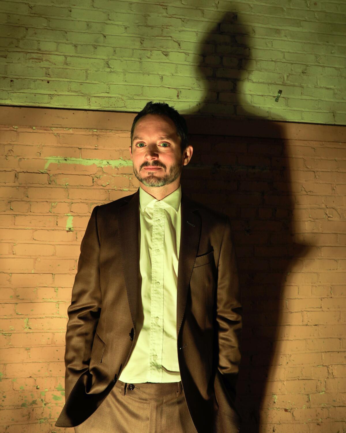 Elijah Wood, wearing a brown suit, stands against a brick wall.