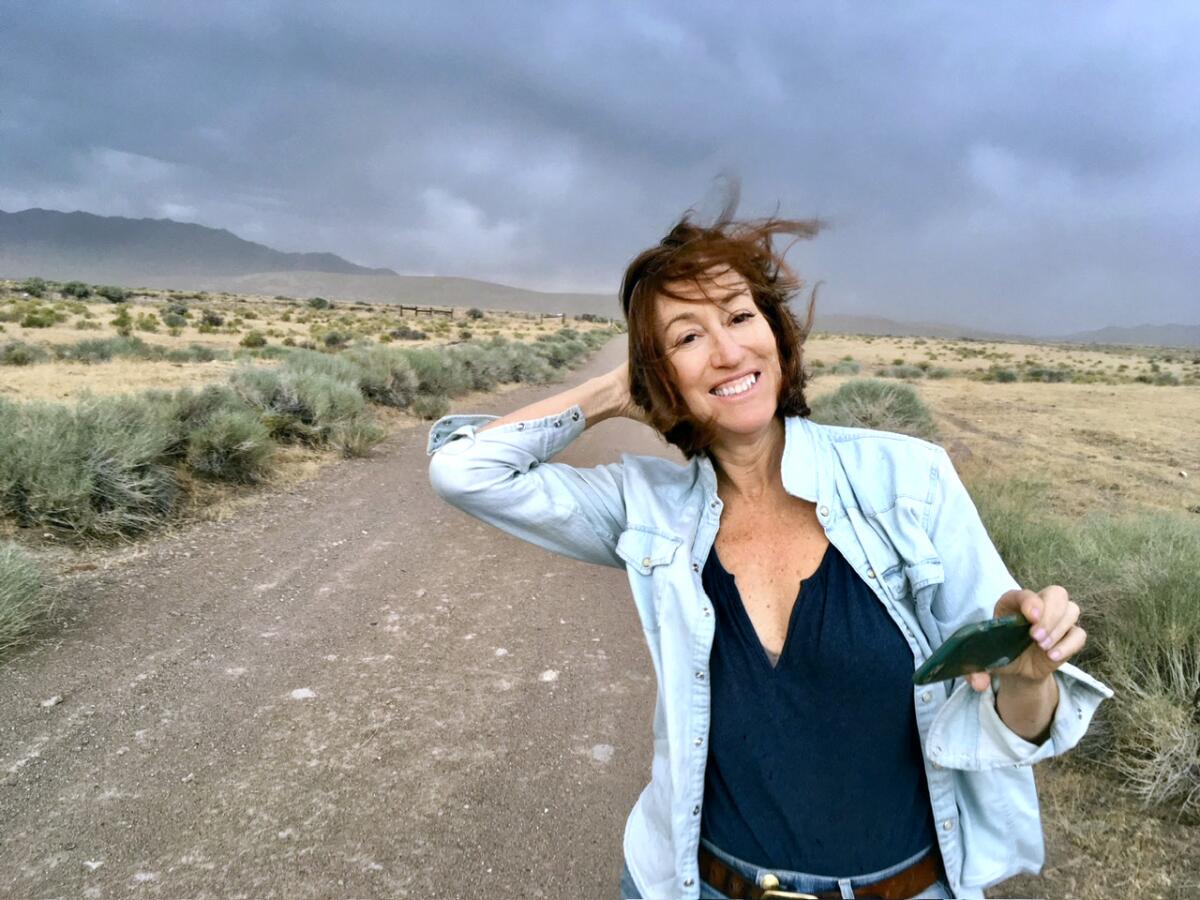 Diana Marcum stands by a dirt road outdoors while wind blows her hair and dark clouds gather in the background.