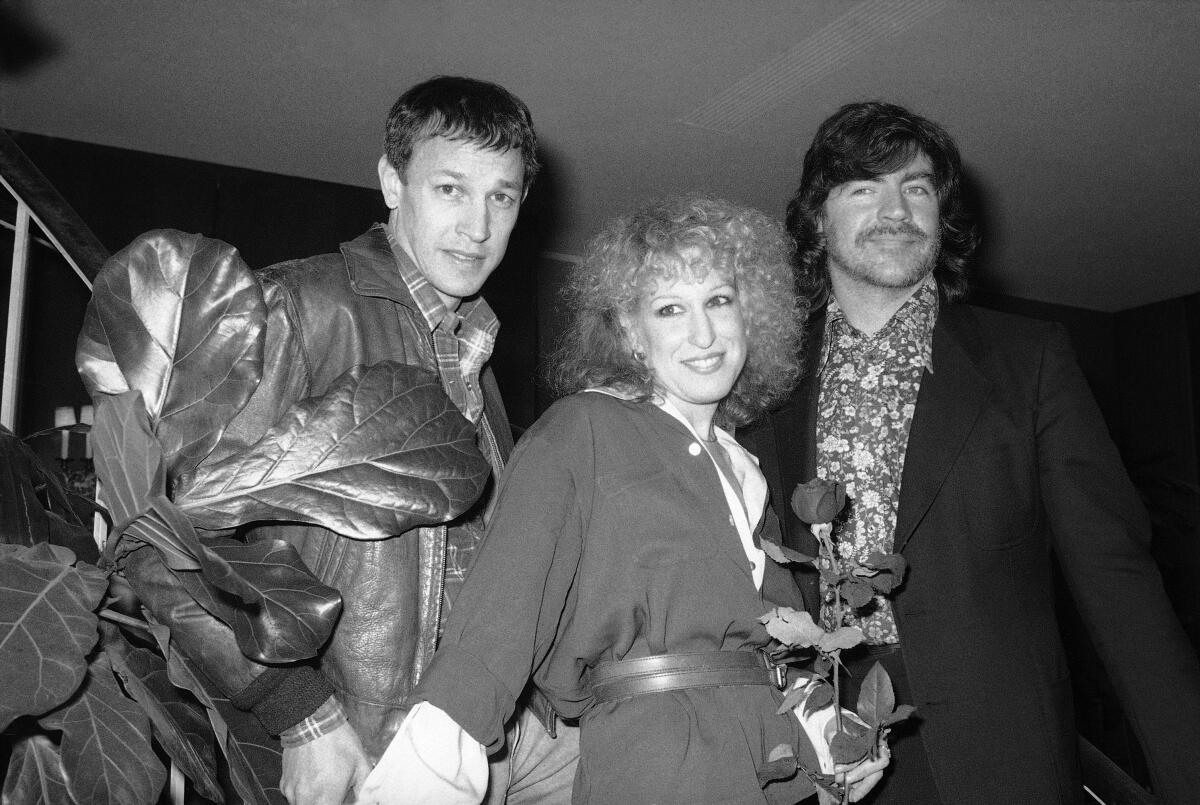 A black-and-white photo of Frederic Forrest, left, Bette Midler and Alan Bates posing on a staircase next to a large plant