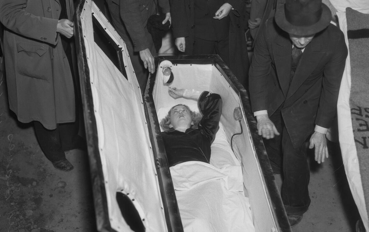 A woman with short blond hair lies inside a coffin and reaches up to a small lightbulb.
