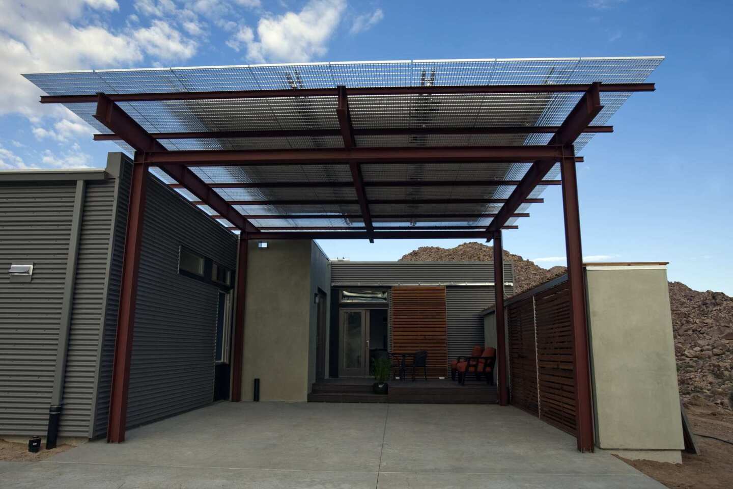 On one side of the house, a carport in front is topped with solar panels. A small deck is tucked by a door to the kitchen. Mechanical systems are hidden behind more cedar-slat panels on the right.
