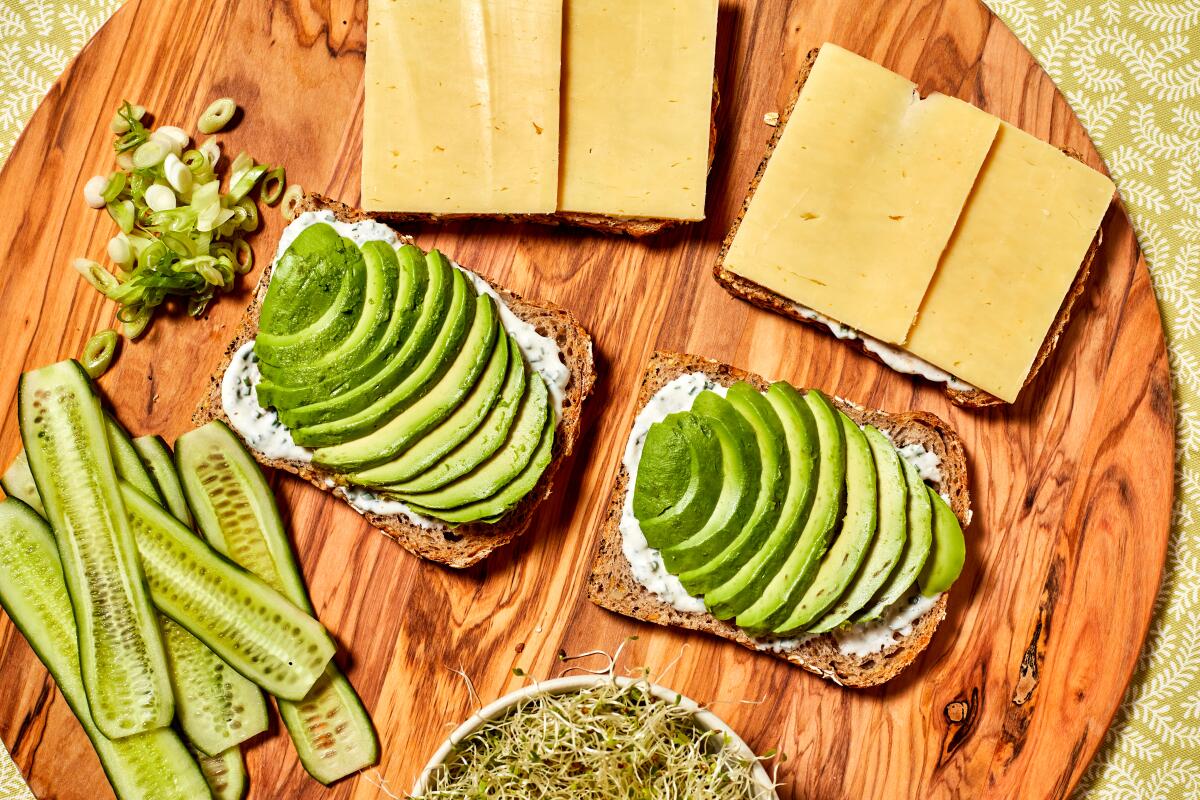 Slices of bread piled with cheese and avocado slices, next to cucumber ribbons and sprouts