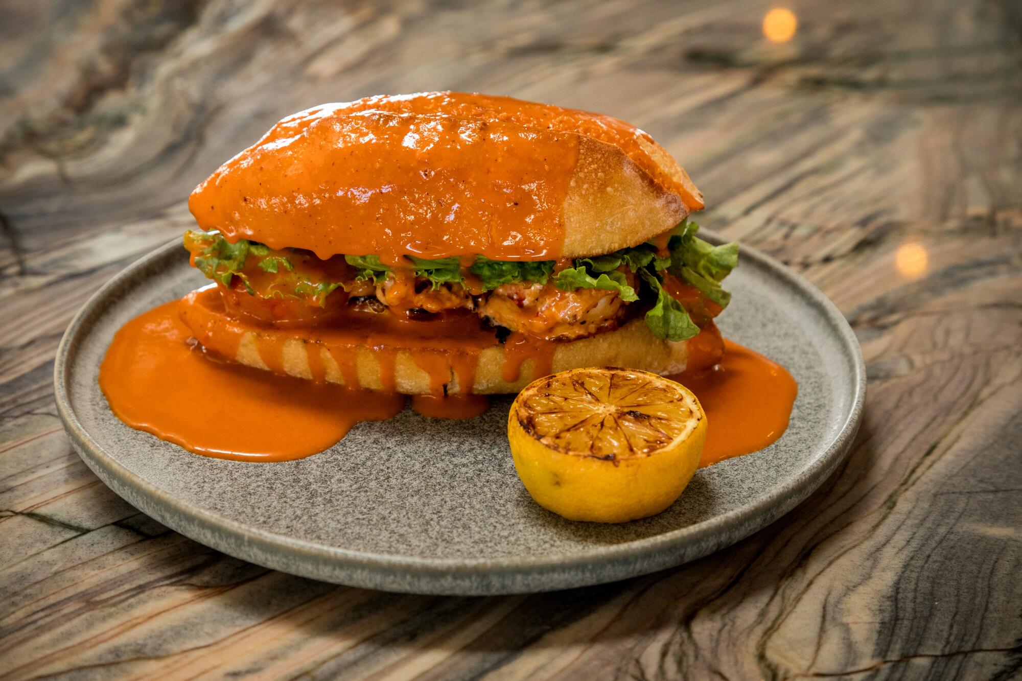 A sandwich on a plate covered in a red-orange sauce, with half a grilled lemon