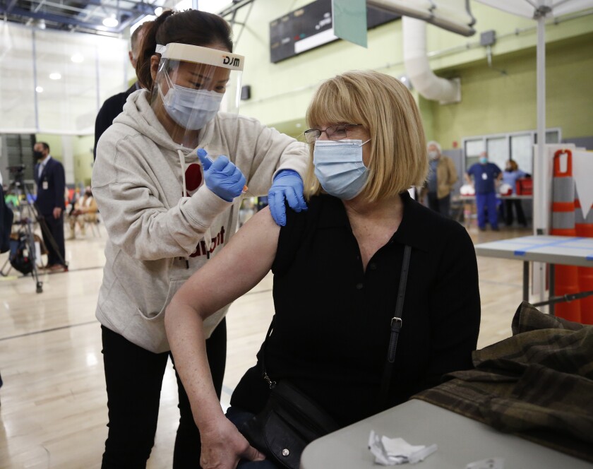 A seated woman with a mask receives a vaccine shot from a nurse