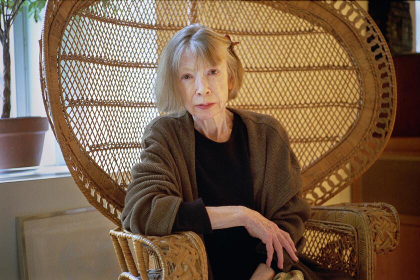 Joan Didion, in a brown sweater, sits in a large, wicker peacock chair.