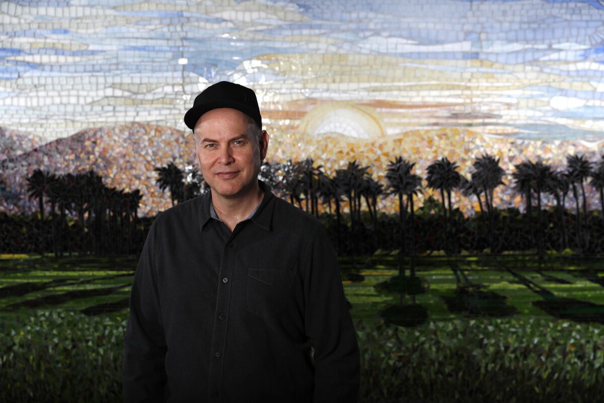 A man in a black shirt and cap stands in front of a mosaic of palm trees and green lawn.