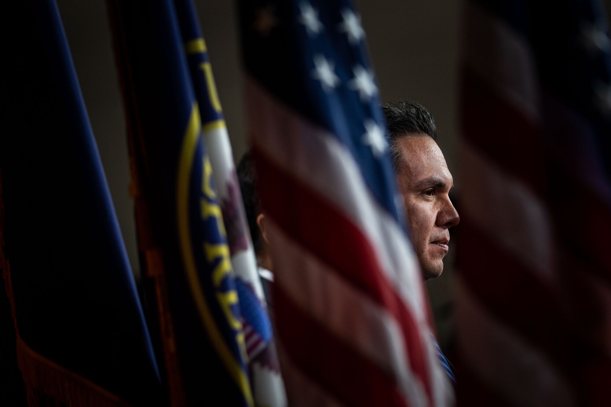 Pete Aguilar is framed head-and-shoulders from a profile, looking toward to right side, standing between flags.